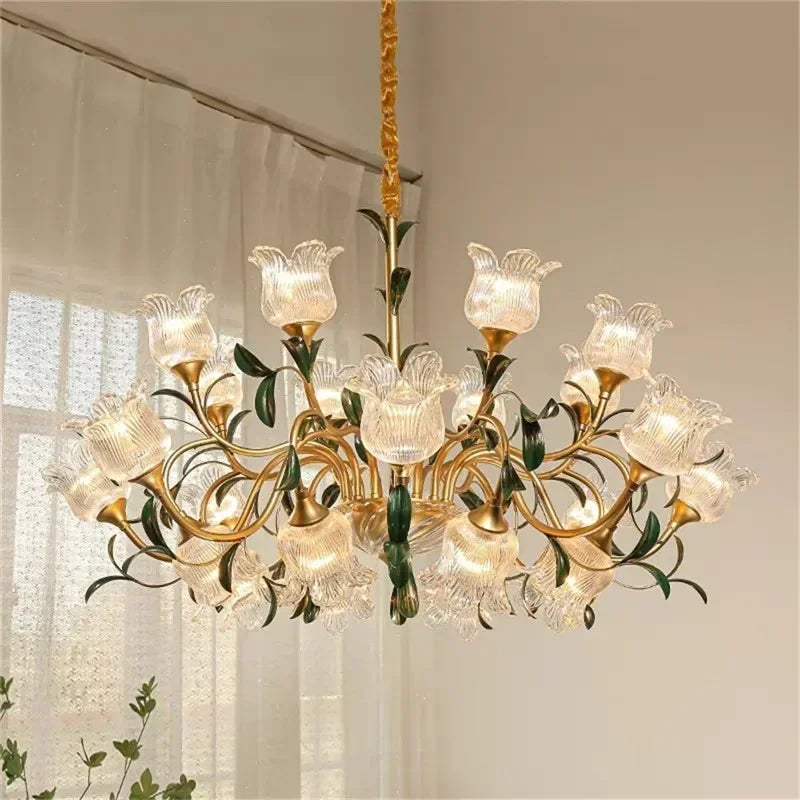 Wide shot of a multi-arm gold chandelier with clear glass floral shades, installed in a softly lit room with curtains in the background.