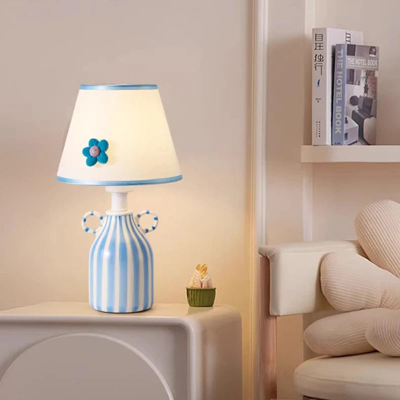 Blue and white striped ceramic table lamp with a scalloped white fabric shade featuring a blue flower accent, placed on a white nightstand next to a shelf with books.