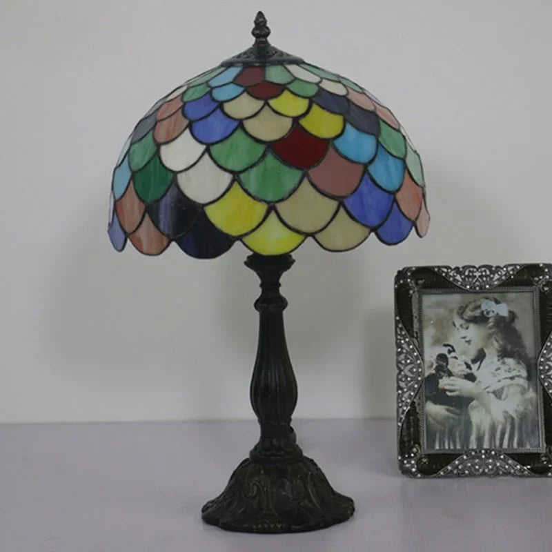 A Tiffany-style table lamp with a multicolored stained glass dome shade and a dark iron base, positioned on a surface.