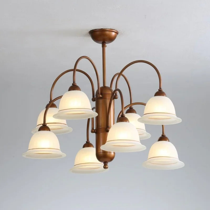 A view of a brushed gold chandelier with eight frosted glass bell-shaped shades, illuminated and casting a warm glow, against a subtly textured ceiling.