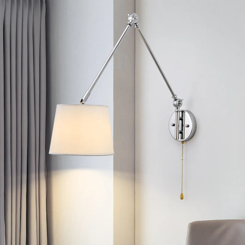 Chrome swing arm wall sconce with a white fabric drum shade and pull chain, shown in a room with grey curtains.