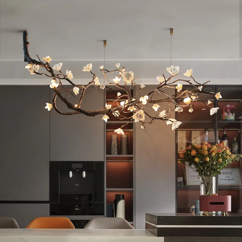 A close-up of a decorative chandelier resembling a tree branch with white flowers, illuminated by warm LED lights, installed in a modern dining area.