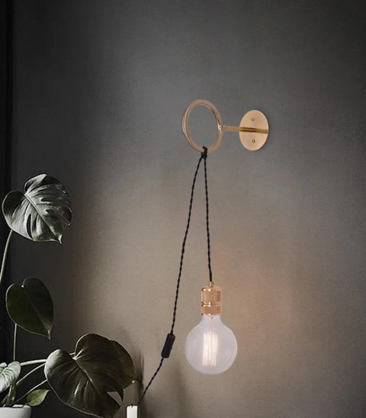 Wall-mounted light fixture with a bulb on a dark wall, next to a plant.