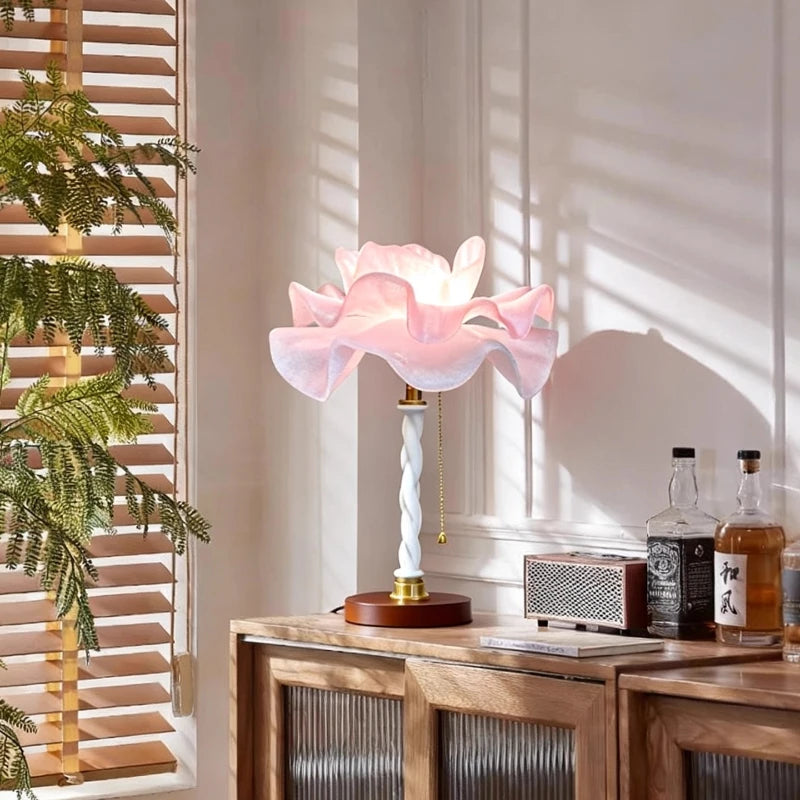 A white and pink petal-shaped table lamp illuminated on a wooden cabinet, with blinds casting shadows in the background.