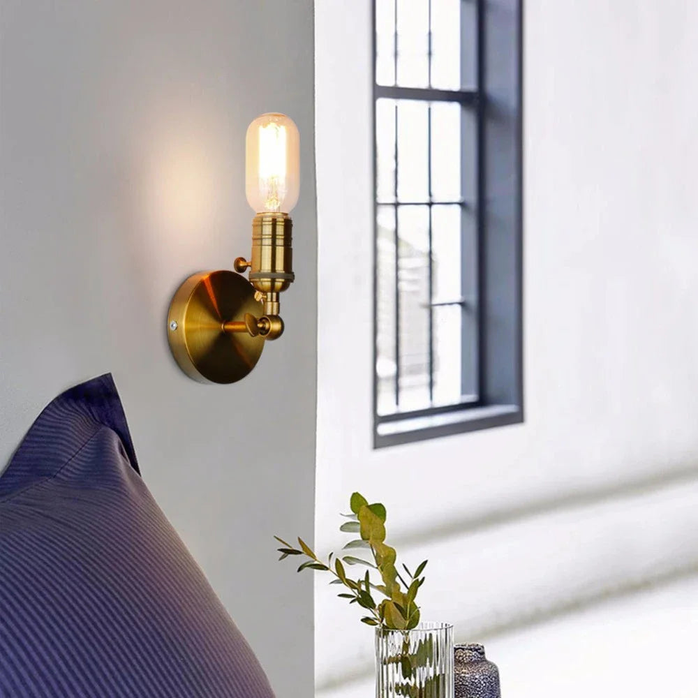 Close-up of a gold industrial-style wall sconce with an adjustable folding arm and exposed Edison-style bulb, mounted on a white wall next to a blue pillow.