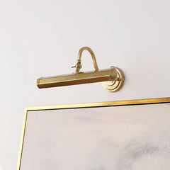 Brushed brass wall sconce with a curved arm and rectangular light head positioned above a framed artwork.