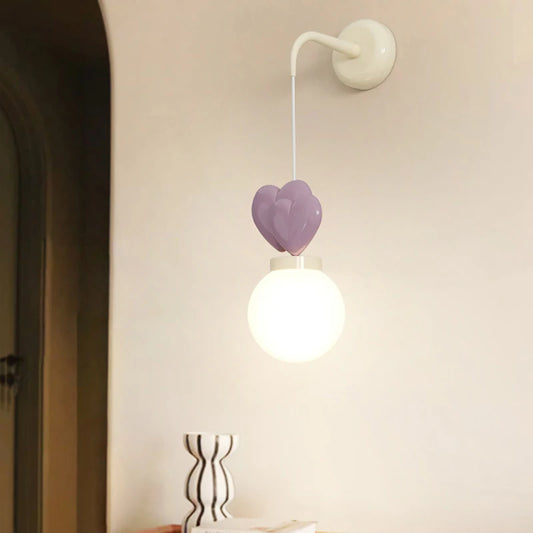 A wall sconce with a white mounting bracket, a white cord, three purple heart-shaped accents, and a white globe light emitting warm light.
