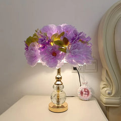 A close-up of a table lamp with a gold crystal base and a vibrant purple floral arrangement on top, emitting a soft light.