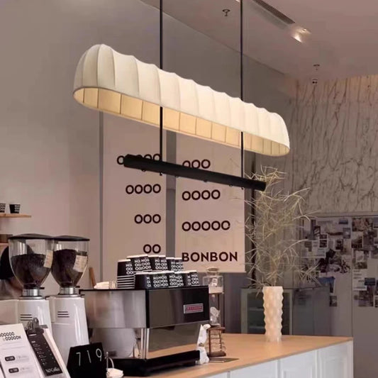 Close-up of a cream fabric tube pendant light with a black bar below, hanging in a cafe setting with menu boards in the background.