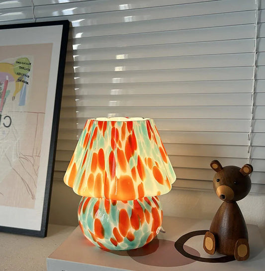 A single mushroom-shaped table lamp with a stained glass shade featuring orange and teal splatters, placed on a book next to a wooden bear figurine.