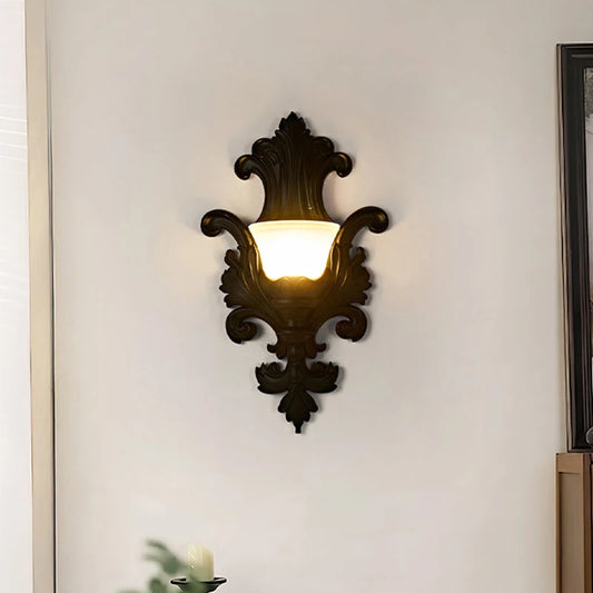 Black ornate resin wall sconce with a frosted glass shade emitting a warm glow, mounted in a hallway.