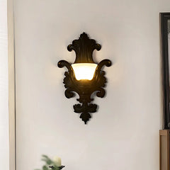 Black ornate resin wall sconce with a frosted glass shade emitting a warm glow, mounted in a hallway.