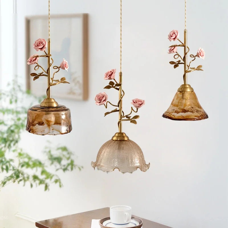 Three different styles of pendant lights, each with a brass stem adorned with pink rose buds and connected to variously shaped amber and clear glass shades.