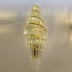 A tall, spiraling gold wall sconce with multiple tiers of clear crystals hanging from gold bands, illuminated against a light-colored wall.