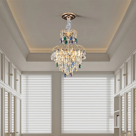 A gold multi-tier crystal chandelier illuminated in a hallway with white built-in cabinets and blinds.