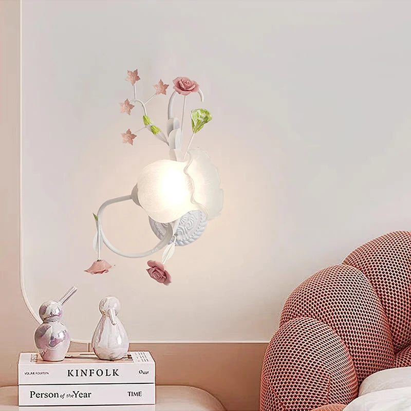 A white floral rose bud wall sconce on a pink wall, with a bedside table featuring decorative items and books below.