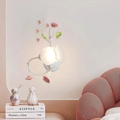 A white floral rose bud wall sconce on a pink wall, with a bedside table featuring decorative items and books below.