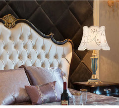 Ornate table lamp with a teal and gold resin base shown in a bedroom setting next to a bed.