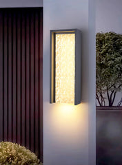 Modern outdoor wall light fixture on a building exterior with plants in the background