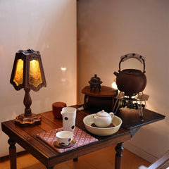 A table lamp with a black walnut and glass shade sits on a wooden table next to tea ceremony items.