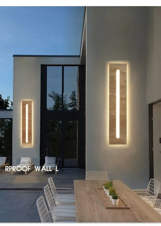 Two modern rectangular wall sconces with natural stone finish and vertical LED lights, illuminating an outdoor patio area with a dining table.