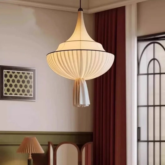 A white fabric pleated pendant light with a black trim, hanging from a white ceiling in a room with a painting on the wall and a red curtain.