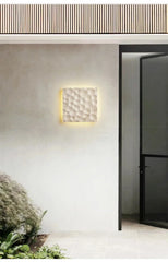 A light stone textured wall sconce mounted on an exterior wall near a modern doorway and lush greenery.