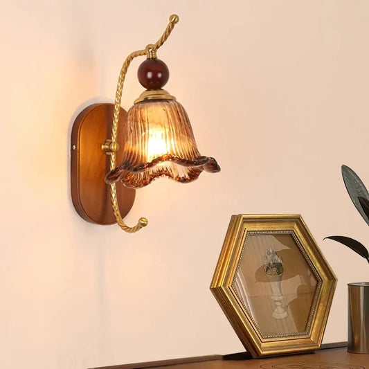 A vintage-style wall sconce with a floral-shaped amber glass shade and a gold arm mounted on a wooden oval backplate.