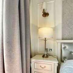 A clear glass cylinder table lamp with a brushed gold base and accents sits on a white nightstand next to a grey curtain and a wall sconce.