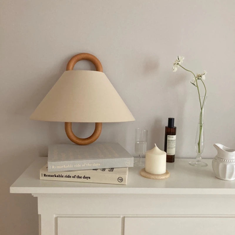 A beige fabric cone shade wall sconce with a natural wood hoop frame mounted on a wall above a shelf with books and decor.