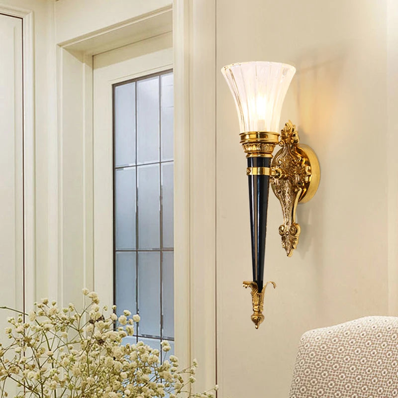 A gold wall sconce with a flared clear glass shade and a dark conical body, mounted on a cream-colored wall next to a window.
