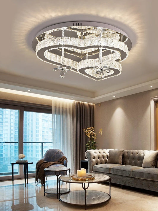 A heart-shaped crystal chandelier with multiple tiers and a polished metal frame, illuminated in a modern living room setting.