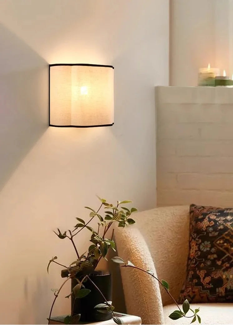 Wall-mounted lamp casting a warm glow in a cozy room with a plant and pillows.