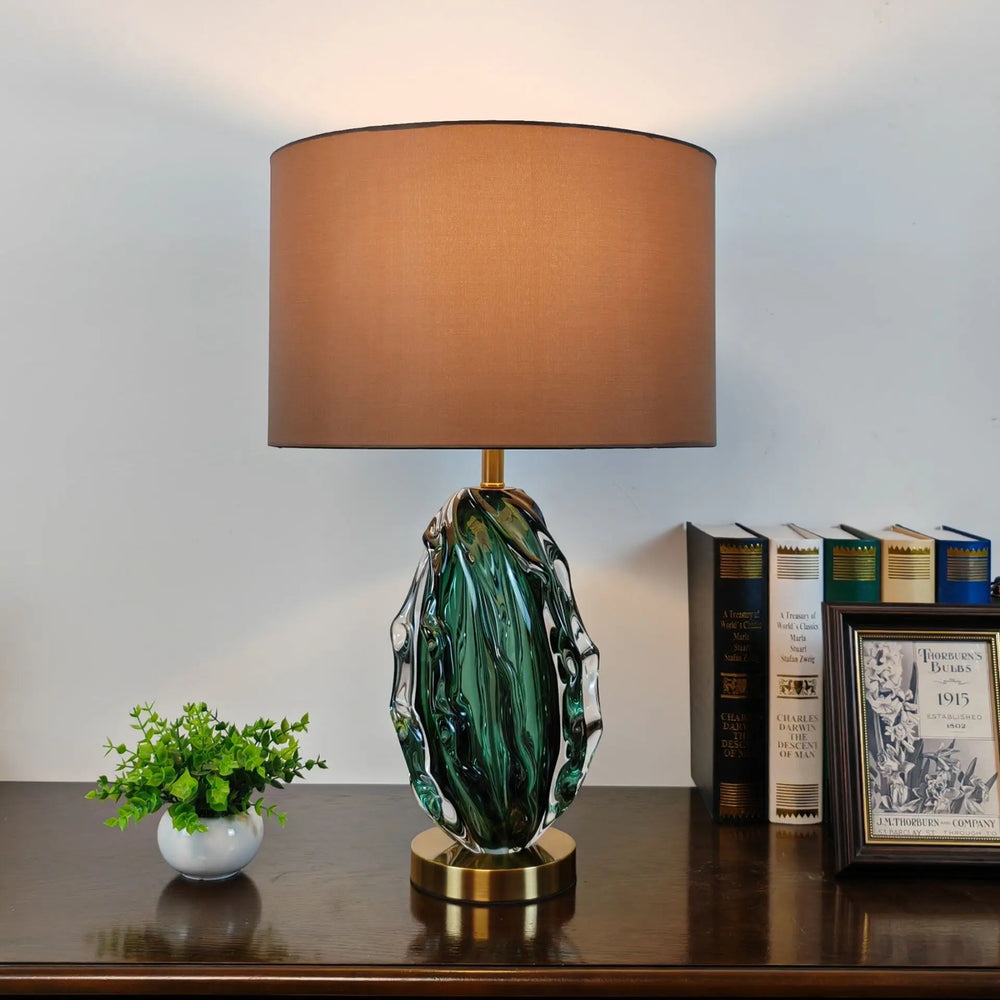 Decorative table lamp with a green base and brown shade on a desk with books and a plant.