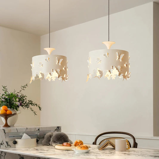 Two cream-colored drum pendant lights with butterfly cutouts hanging above a kitchen island. One light is in focus, showing the detailed design.