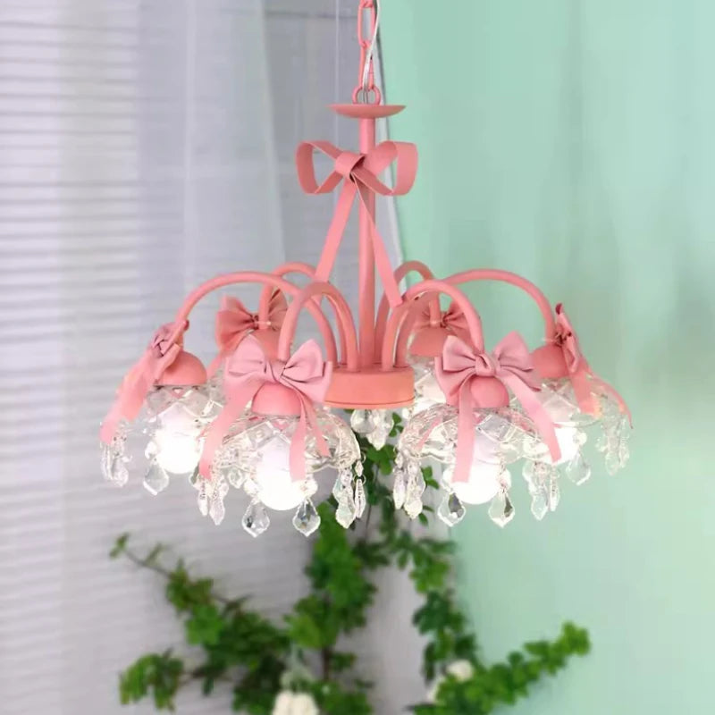 Front view of a pink multi-arm chandelier featuring pink bows, crystal pendants, and clear glass shades, with greenery in the background.