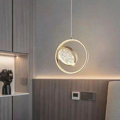 A gold ring pendant light with a crystal diffuser hanging near a modern wood-paneled wall and shelf.