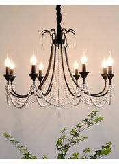 View of a black, multi-arm chandelier with pearl beads and crystal accents, partially framed by green foliage against a light-colored wall.