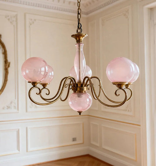 Close-up of a gold pendant light with pink glass globes and ornate arms, mounted in a room with ornate wall paneling.