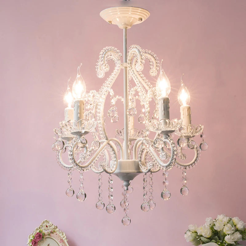 A white candelabra-style chandelier with intricate scrollwork and clear crystal accents, set against a pink background.