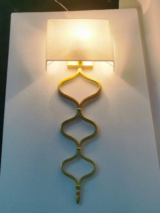 A gold wall sconce with a quatrefoil chain design and fabric shade illuminated against a textured white background.