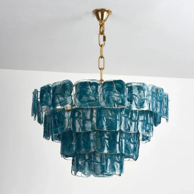 A multi-tiered chandelier with square, textured blue glass panels suspended from a gold chain and frame, against a white ceiling and wall.