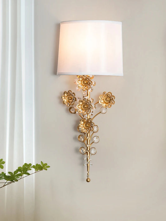 A gold floral crystal wall sconce with a white fabric shade, positioned next to a window with sheer curtains.