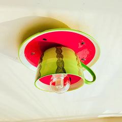 Close-up of a whimsical ceiling light designed to look like a slice of watermelon with a green rind and red interior, featuring a green ceramic coffee cup hanging as the main body, and a visible LED bulb.