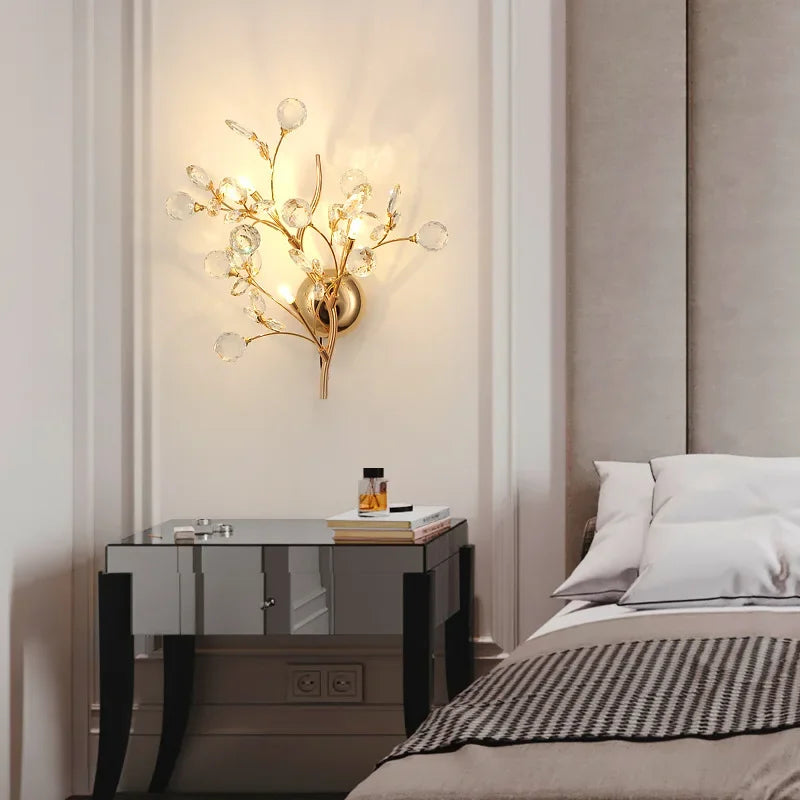 A gold wall sconce with clear crystal accents, emitting light against a cream-colored wall in a bedroom setting.