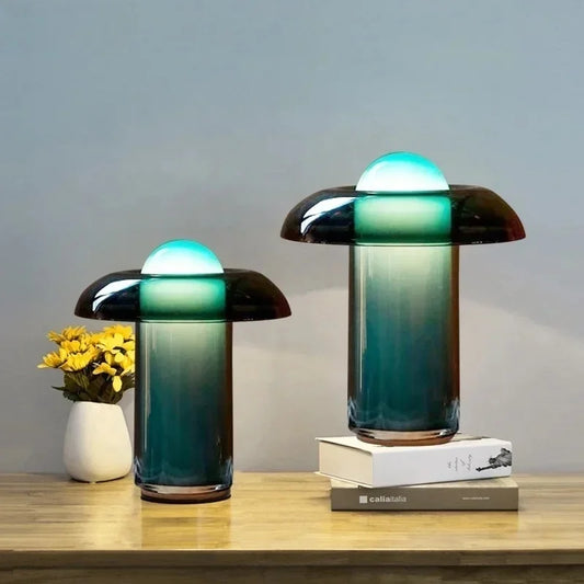 Two glass mushroom table lamps with gradient shades, one on a wooden surface with books and a vase of yellow flowers, the other on a concrete block.