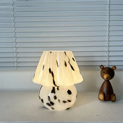 A mushroom-shaped table lamp with a white stained glass shade speckled with black, positioned on a surface next to a wooden bear.