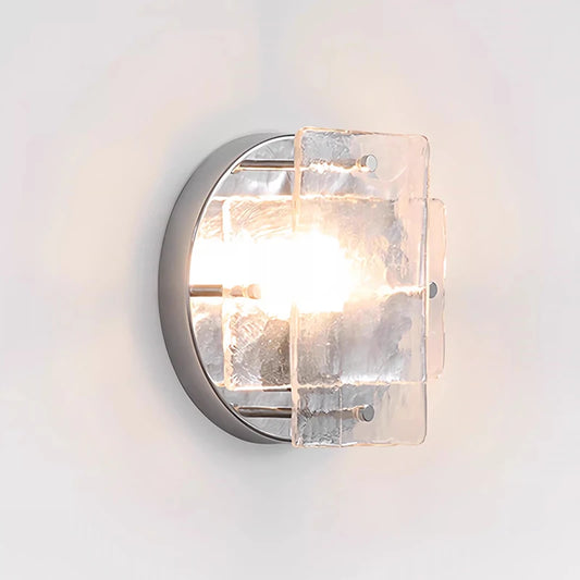 Close-up of a modern silver wall sconce with textured clear glass panels emitting light against a white wall.