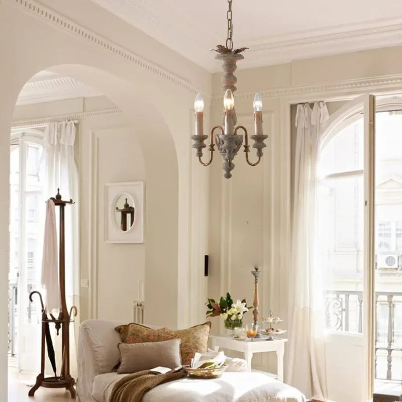 Distressed wood candelabra pendant light hanging in a bright, airy living room with large windows, sheer curtains, and a chaise lounge.
