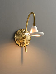 A gold floral wall sconce with a white ceramic shade and curved arm, mounted on a grey wall, emitting warm light.